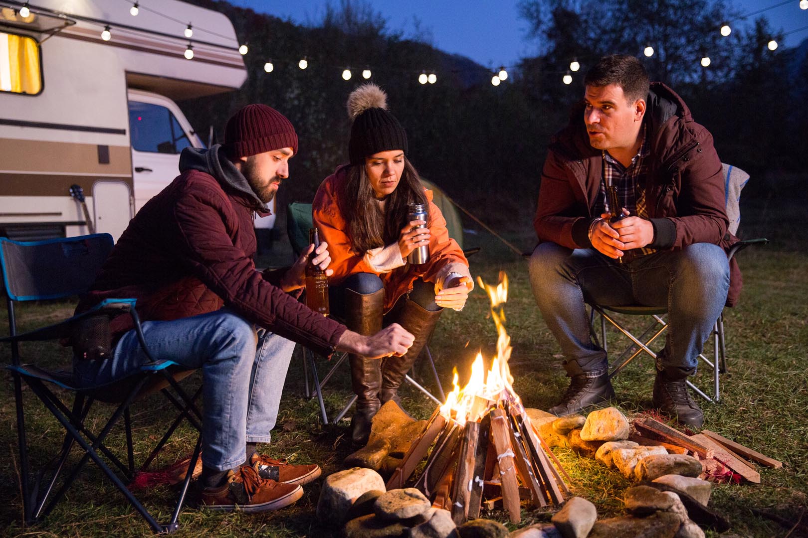 Campers relaxing together around camp fire drinking beer retro camper van background