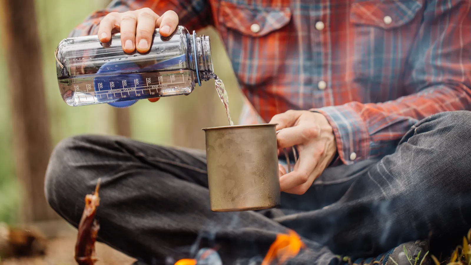 Pouring stored water from a bottle into a metal mug beside the campfire