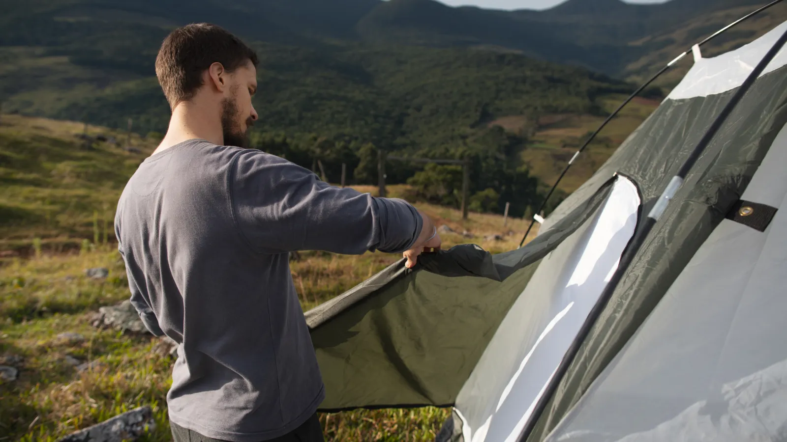 Camper pitching a tent on a grassy hillside on a summer morning